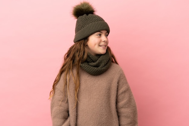 Bambina con cappello invernale isolato su sfondo rosa guardando di lato e sorridente