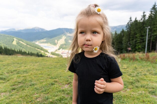 Bambina con fiore in labbra che distoglie lo sguardo in piedi sul picco con vista sulle montagne dietro