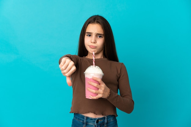 Bambina con frappè alla fragola isolato su sfondo blu che mostra il pollice verso il basso con espressione negativa