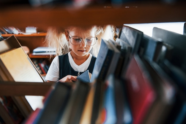 Bambina con gli occhiali alla ricerca di un libro in biblioteca