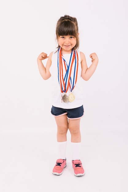 Bambina con i capelli castani con medaglie di campioni sportivi sul collo che fanno il simbolo della forza. Sport e concetto di vittoria