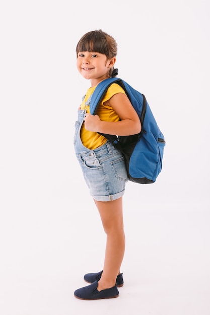 Bambina con i capelli neri vestita con una salopette di jeans e una t-shirt blu, con uno zaino pronto per tornare a scuola, su un fianco, su sfondo bianco