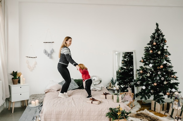 Bambina con la mamma a letto in camera da letto vicino all'albero di Natale Felice Anno Nuovo e Buon Natale