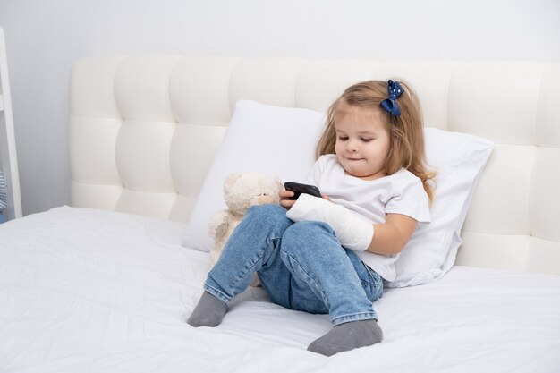 Bambina con la mano in cast seduto nel letto utilizzando smartphone, guardando cartoni animati o video educativi.