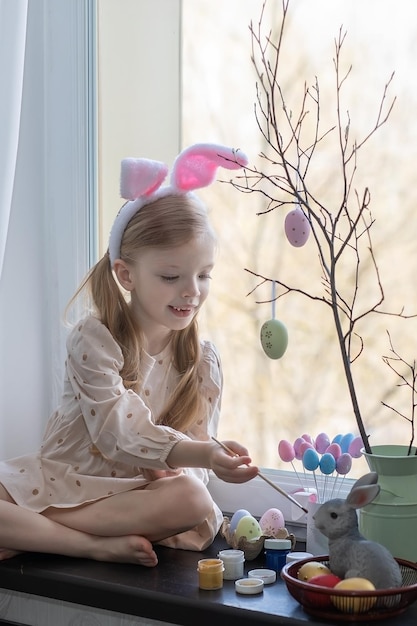 bambina con le orecchie di coniglio in testa sta dipingendo le uova per un cesto di Pasqua sul davanzale della finestra