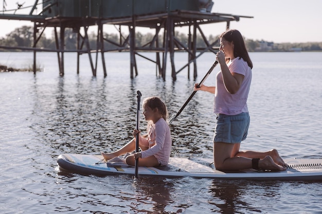 Bambina con madre sup che si imbarcano insieme su un sup di giorno con casa sull'acqua sullo sfondo Stile di vita attivo Tempo insieme Insegnare ai bambini a fare sport fin dalla tenera età