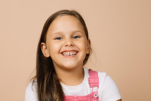 Bambina con occhi luminosi e luminosi e un sorriso beato eccitato con i denti che guardano la telecamera che indossa una tuta rosa brillante e una maglietta bianca su sfondo beige