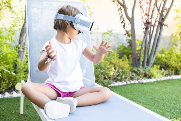 Bambina con occhiali per realtà virtuale guardando a destra cercando di toccare qualcosa in modo virtuale seduto su un lettino nel giardino di casa sua Metaverse VR gioco concetto digitale