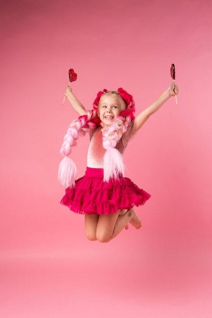 bambina con trecce di kanekalon rosa detiene una lecca-lecca a forma di cuore su uno sfondo rosa