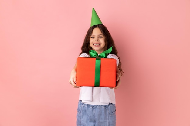 Bambina con umore festivo in piedi nel cono di festa e dando una scatola regalo per festeggiare il compleanno