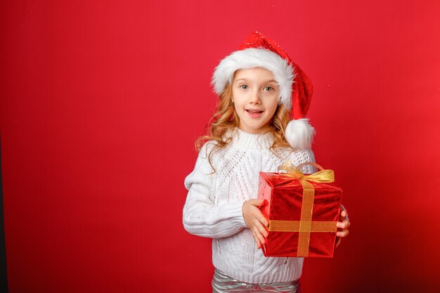 Bambina con un cappello da Babbo Natale su uno sfondo rosso con una torta e un regalo