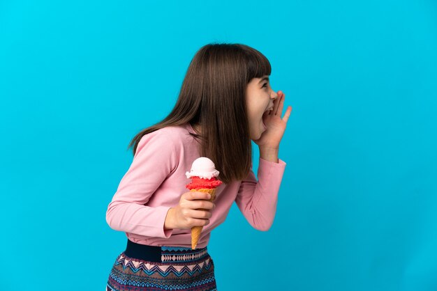 Bambina con un gelato alla cornetta isolato su sfondo blu che grida con la bocca spalancata di lato