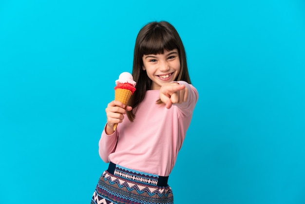 Bambina con un gelato alla cornetta isolato su sfondo blu che punta davanti con espressione felice