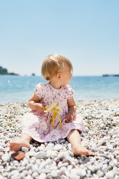 Bambina con una banana in mano si siede su una spiaggia di ciottoli girando la testa di lato