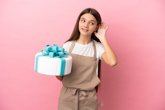 Bambina con una grande torta su sfondo rosa isolato che ascolta qualcosa mettendo la mano sull'orecchio