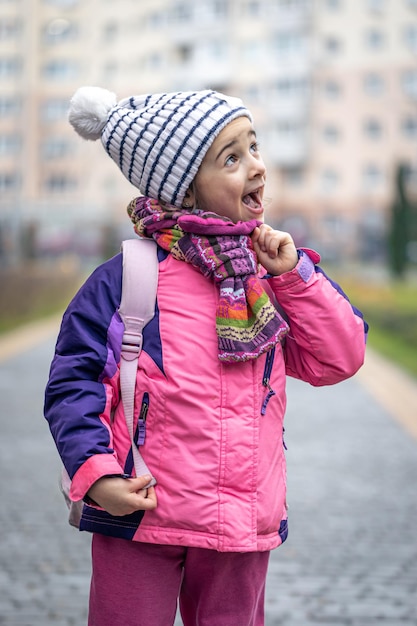 Bambina con uno zaino in giacca e cappello vicino alla scuola