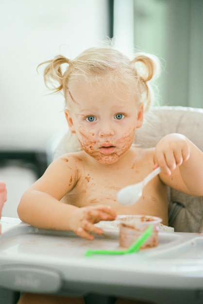 Bambina dai capelli biondi con la faccia disordinata quando si mangia un pasto seduto nel seggiolone