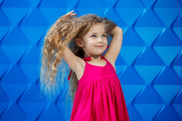 Bambina dai capelli biondi in un vestito rosa su uno sfondo blu che balla e ride, emozioni di gioia per bambini brillanti, infanzia felice