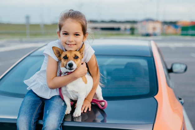 Bambina dall'aspetto accattivante, abbraccia il suo animale domestico preferito, viaggia insieme ai genitori in auto, si siede al bagagliaio, posa per fare foto. Concetto di bambini, animali, riposo e trasporto