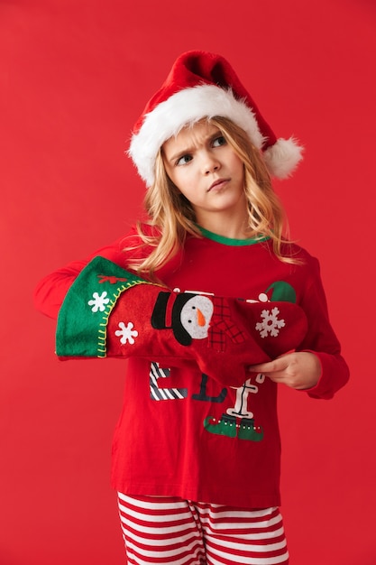 Bambina delusa che indossa il costume di Natale in piedi isolato, prendendo i regali da un calzino di Natale