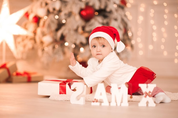 Bambina di 1 anno che indossa un abito di babbo natale che striscia sull'albero di Natale con luci. Vacanze. Infanzia.