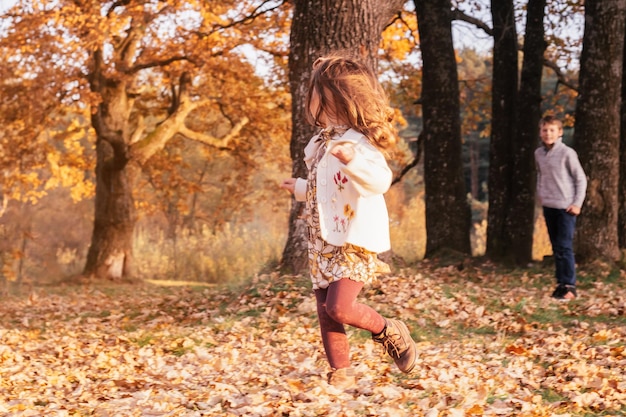 Bambina di 34 anni corre dal fratello ragazzo 910 anni su foglie di autunno all'aperto vicino a spessi tronchi di quercia nel parco