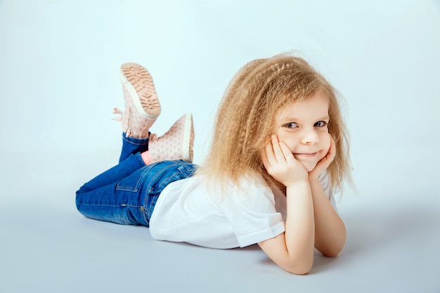 Bambina di 4 anni con i capelli ricci che indossa camicia bianca, blue jeans disteso sul pavimento, sorridendo e guardando, mani che tengono la testa