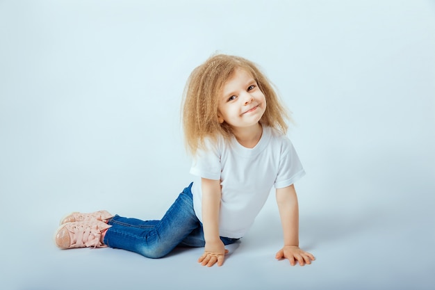Bambina di 4 anni con i capelli ricci che indossa camicia bianca, blue jeans, stivali rosa seduto sul pavimento, sorridendo e guardando la telecamera.