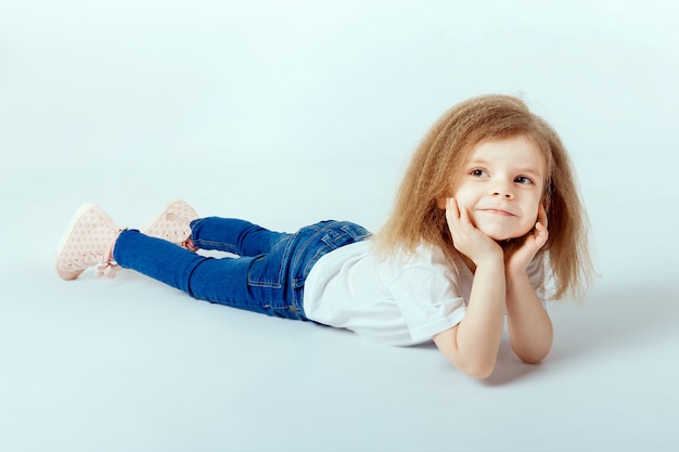 Bambina di 4 anni con i capelli ricci che indossa una camicia bianca, blue jeans disteso sul pavimento, sorridendo e guardando la telecamera, mani che tengono la testa