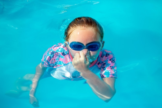 Bambina di 8 anni, in costume da bagno luminoso e occhiali blu, nuota, si tuffa, si tuffa sott'acqua in una piscina all'aperto al sole con acqua blu