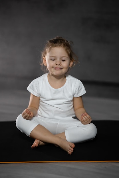 Bambina di tre anni che medita in una postura di loto su uno sfondo grigio in una stanza buia
