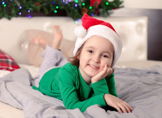 Bambina divertente berretto da Babbo Natale e pigiama natalizio giacciono sul letto a casa