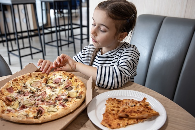 Bambina divertente che mangia pizza in una scatola di cartone per pranzo.