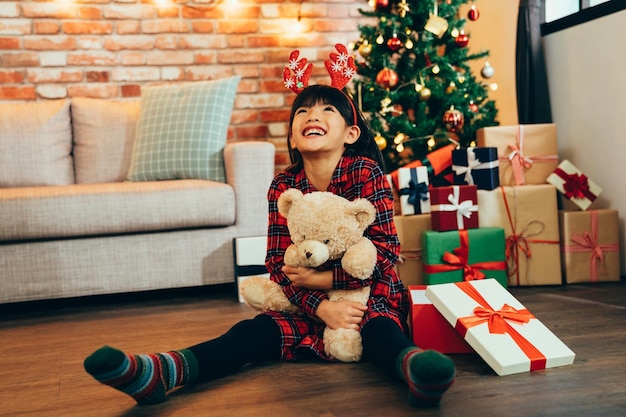 Bambina dolce che abbraccia l'orsacchiotto guardando il cielo sorridente seduto sul pavimento di legno. infanzia attraente ha ricevuto un regalo in vendita di Natale a casa. capretto con renna soddisfatto del presente.