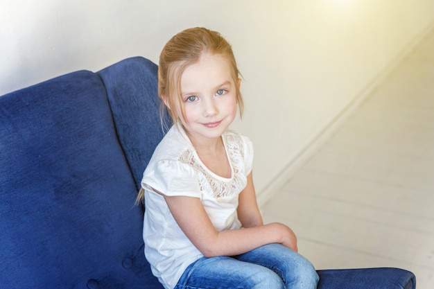 Bambina dolce in jeans e maglietta bianca a casa che si siede sulla sedia blu accogliente moderna che si rilassa nel salone bianco. La gioventù degli scolari dell'infanzia rilassa il concetto