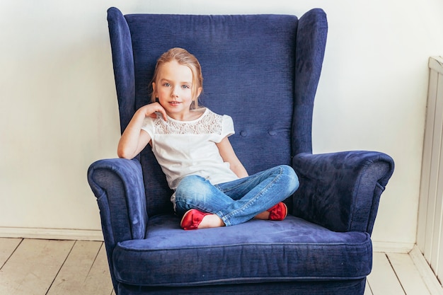 Bambina dolce in jeans e maglietta bianca che si siedono sulla sedia blu accogliente moderna che si rilassa a casa