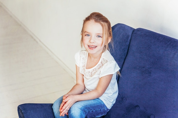 Bambina dolce in jeans e maglietta bianca che si siedono sulla sedia blu accogliente moderna che si rilassa a casa