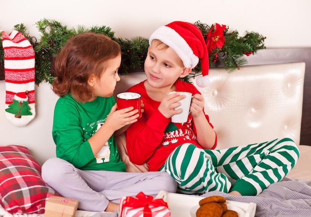 Bambina e bambino in berretto da Babbo Natale e pigiama di Natale che bevono cacao
