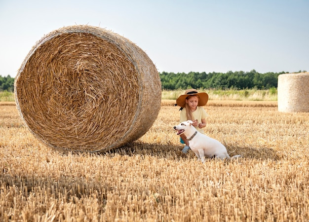 Bambina e cane che si divertono in un campo di grano in una giornata estiva. Bambino che gioca al campo di balle di fieno durante il periodo del raccolto.