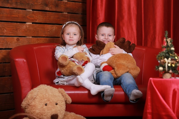 Bambina e ragazzo sul divano in studio di Natale nei colori rossi. albero di natale e orsacchiotto sul muro.