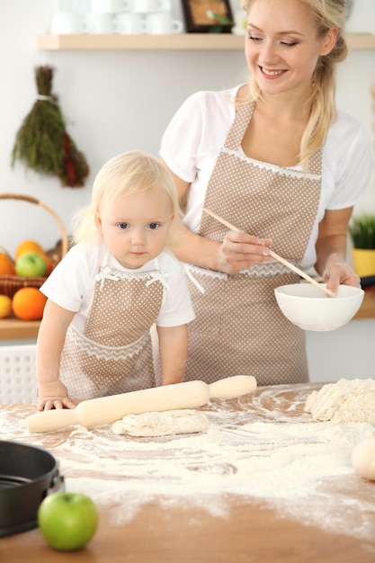 Bambina e sua mamma bionda in grembiuli beige che giocano e ridono mentre impastano la pasta in cucina. Pasticceria fatta in casa per pane, pizza o biscotti da forno. Divertimento in famiglia e concetto di cucina.