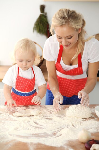 Bambina e sua mamma bionda in grembiuli rossi che giocano e ridono mentre impastano la pasta in cucina. Pasticceria fatta in casa per pane, pizza o biscotti da forno. Divertimento in famiglia e concetto di cucina.