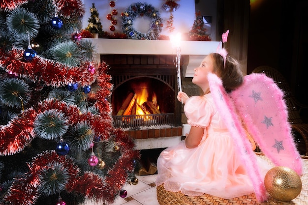 Bambina fata con la bacchetta magica vicino a un camino di albero di Natale sullo sfondo