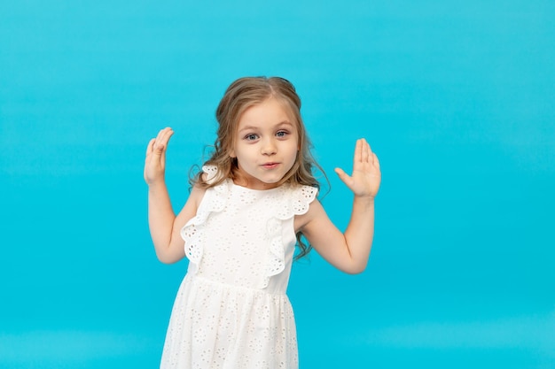 Bambina felice carina in un vestito bianco di cotone su sfondo blu in studio alzò le mani e scherzava in un posto per il testo