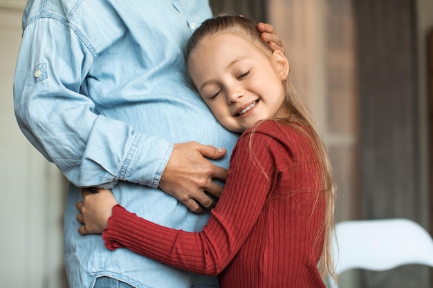 Bambina felice che abbraccia la pancia incinta della madre e sorride con gli occhi chiusi bambina che si appoggia sulla pancia della mamma