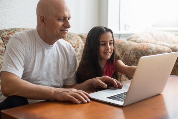 Bambina felice che abbraccia nonno sorridente che si siede sul sofà con il computer portatile.