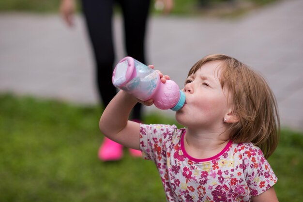 bambina felice che beve acqua fresca da una bottiglia mentre corre al parco cittadino