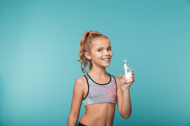 Bambina felice che indossa abbigliamento sportivo acqua potabile da una bottiglia isolata sopra la parete blu