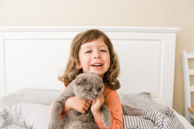 Bambina felice con gatto a letto Bambina che sorride e si diverte con il gatto scozzese