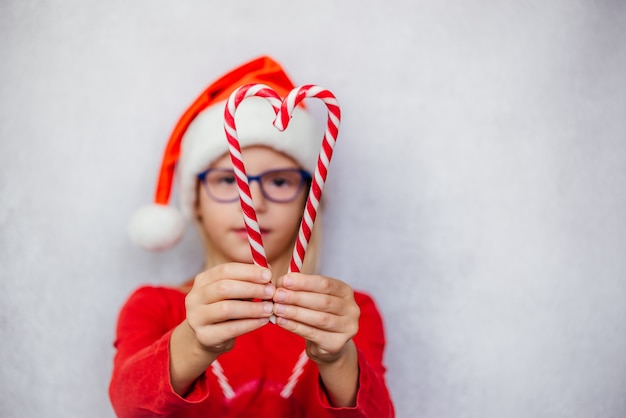 Bambina felice con gli occhiali e il cappello di Babbo Natale a forma di cuore con bastoncini di zucchero, oftalmologia del bambino di Natale